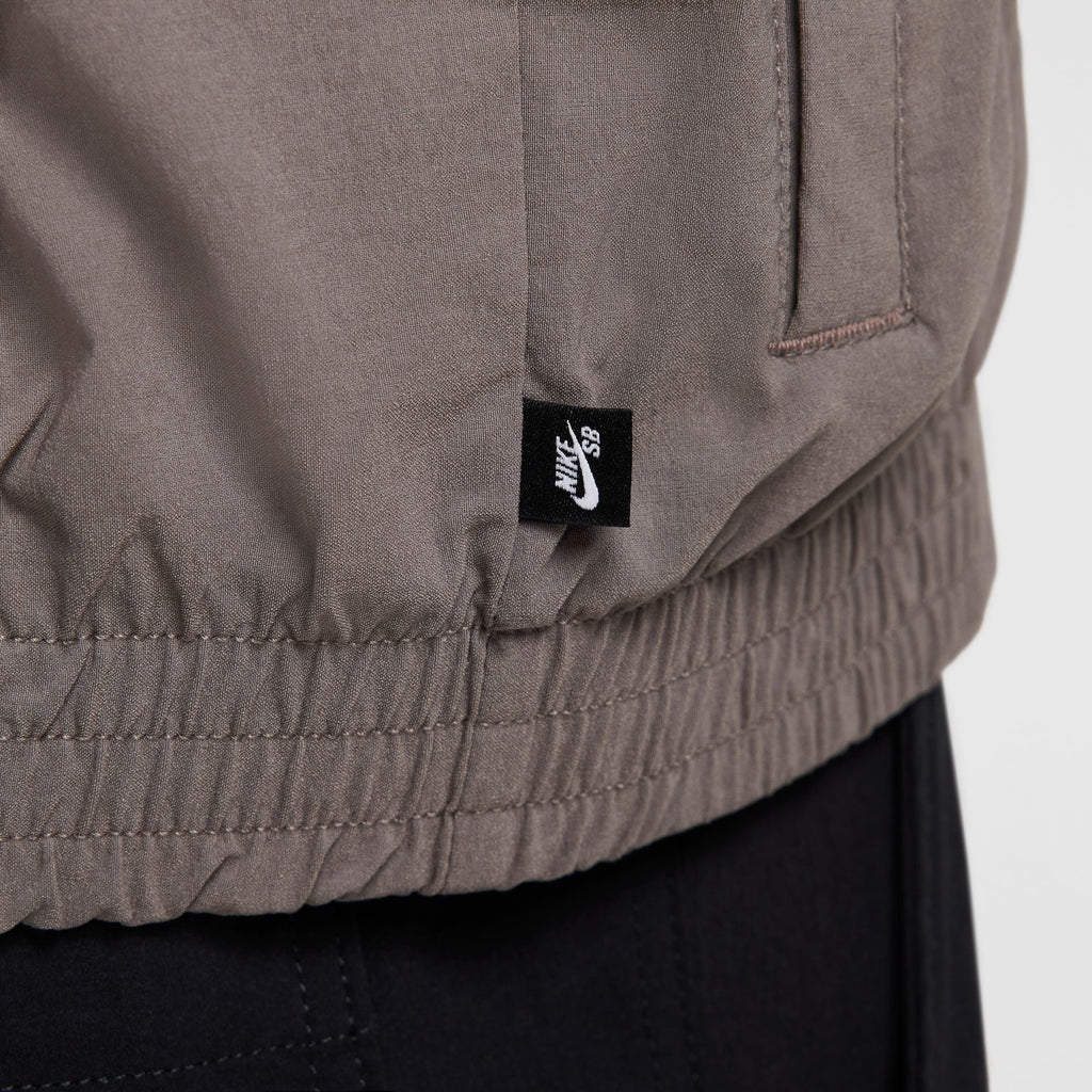 Close-up of the NIKE SB FULL-ZIP WOVEN SKATE JACKET in Sequoia/Cave Stone/White with a black fabric Nike SB tag on the side, featuring an elasticized hem similar to a Skate Hoodie and part of a pocket.