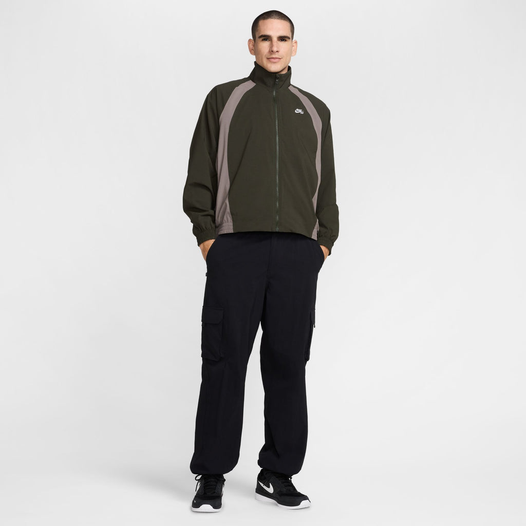 A person wearing a NIKE SB Full-Zip Woven Skate Jacket in Sequoia/Cave Stone/White, paired with black cargo pants and black sneakers, stands against a plain white background.