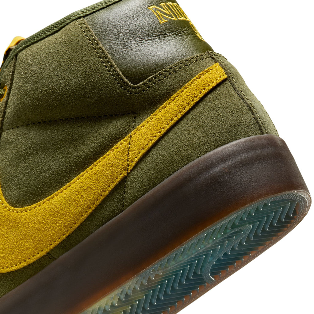 Close-up of a green and yellow sneaker with a brown sole and visible stitching, ideal for skateboarding. The shoe showcases the iconic swoosh logo, is part of the NIKE SB X ANTIHERO BLAZER GT MID collection in Rough Green/Amarillo-Rough Green, and features a textured sole pattern.