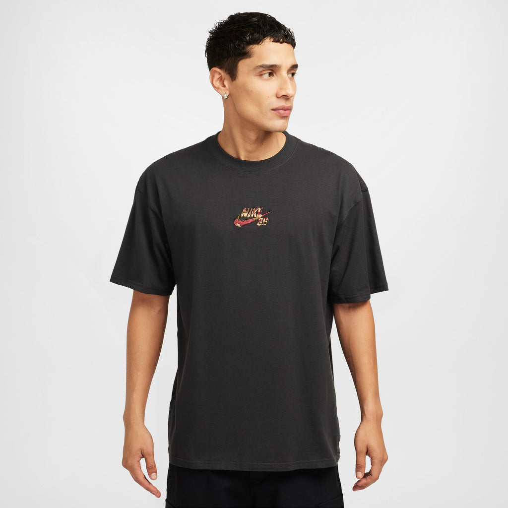 A person wearing a NIKE SB Camo Centered Logo Max90 Tee in Off Noir features a small camo logo on the chest, stands in front of a plain background, looking to the side.