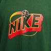 Close-up of a green NIKE SB SONICS MAX90 TEE FIR by Nike, featuring a colorful "Nike SB" logo in red, yellow, and black with stylized lettering that evokes the vibrant spirit once captured by the Seattle SuperSonics.