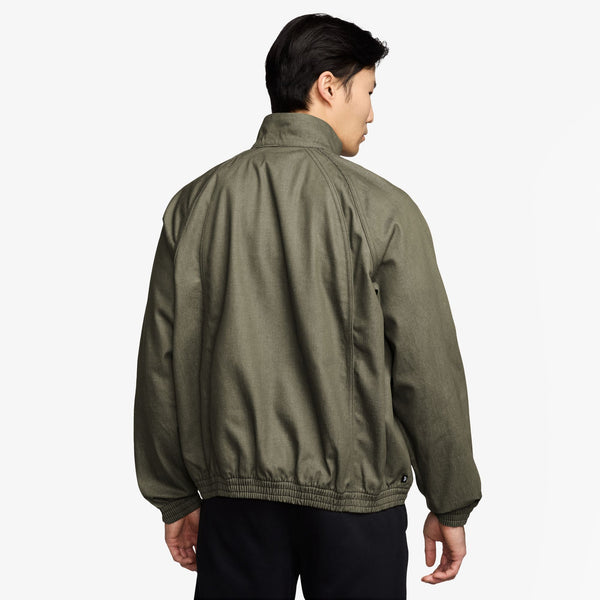 A person is shown from the back wearing a NIKE SB DENIM FULL-ZIP TRACK JACKET in medium olive/white and black pants, set against a plain white background.