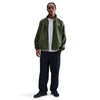 A person stands facing forward, wearing the NIKE SB DENIM FULL-ZIP TRACK JACKET in Medium Olive/White by Nike, a white T-shirt, black cargo pants, and black Nike SB shoes against a plain white background.