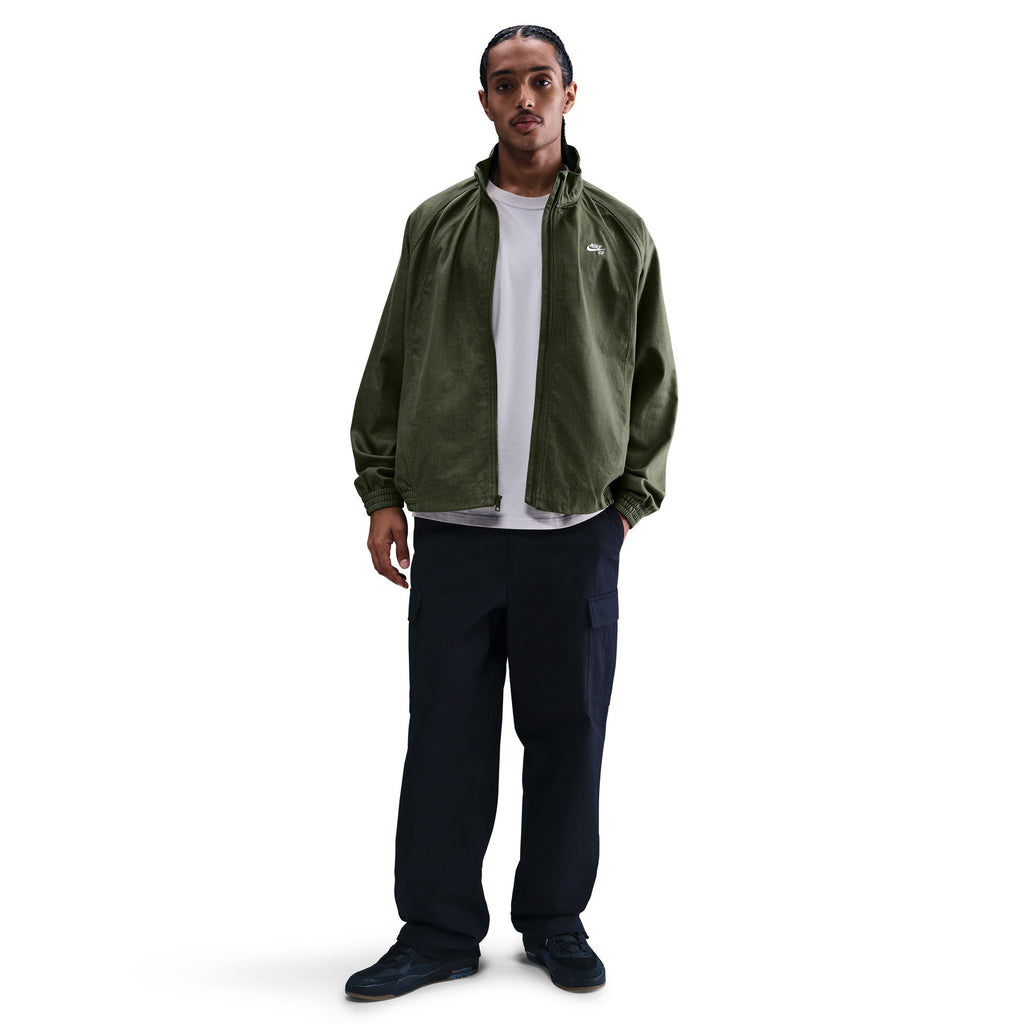 A person stands facing forward, wearing the NIKE SB DENIM FULL-ZIP TRACK JACKET in Medium Olive/White by Nike, a white T-shirt, black cargo pants, and black Nike SB shoes against a plain white background.