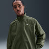 A man faces the camera wearing a nike NIKE SB Denim Full-Zip Track Jacket in Medium Olive/White, featuring a high collar, set against a gradient gray background.