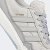 Close-up of a light gray ADIDAS skate shoe with white stripes, light blue sole, and "TYSHAWN" embossed in gold. These TYSHAWN 2 LIGHT ONYX/CLEAR GRANITE shoes offer impact cushioning for style and comfort.