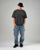 A man in the WARSAW LTD CHAPTER MID | BLACK / BLACK stands hands-in-pockets, wearing a black cap, dark tee, and paint-splattered blue jeans against a grey background—showcasing comfort and durability.