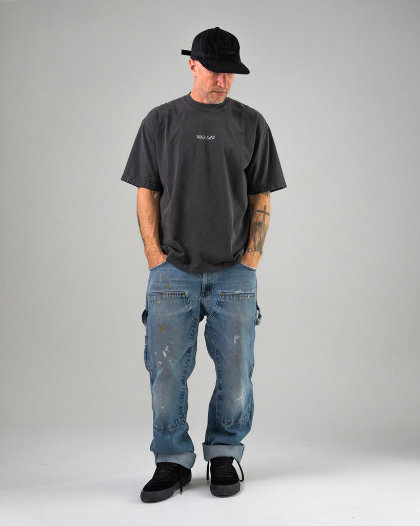 A man in the WARSAW LTD CHAPTER MID | BLACK / BLACK stands hands-in-pockets, wearing a black cap, dark tee, and paint-splattered blue jeans against a grey background—showcasing comfort and durability.