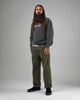 A man with long hair and a beard stands against a gray background, wearing a dark sweatshirt, olive green pants, and black WARSAW LTD STRANGER SLIP-ON shoes—skate-inspired footwear in genuine pig suede.