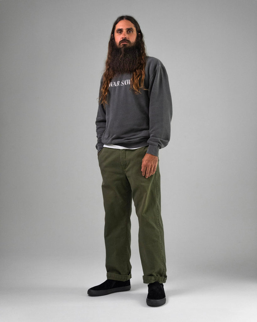 A man with long hair and a beard stands against a gray background, wearing a dark sweatshirt, olive green pants, and black WARSAW LTD STRANGER SLIP-ON shoes—skate-inspired footwear in genuine pig suede.