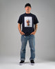 A man stands facing forward with hands in his pockets, wearing WARSAW LTD CHAPTER | BLACK / WHITE SYNTHETIC footwear, a black beanie, navy flower vase print T-shirt, and blue jeans against a plain background.