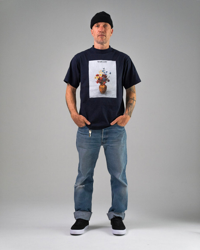 A man stands facing forward with hands in his pockets, wearing WARSAW LTD CHAPTER | BLACK / WHITE SYNTHETIC footwear, a black beanie, navy flower vase print T-shirt, and blue jeans against a plain background.