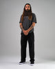 A man with long hair and a beard stands against a plain gray background, wearing a dark T-shirt, black pants, and WARSAW LTD STRANGER SLIP-ON shoes in black/white synthetic with a minimalistic skate-inspired silhouette.