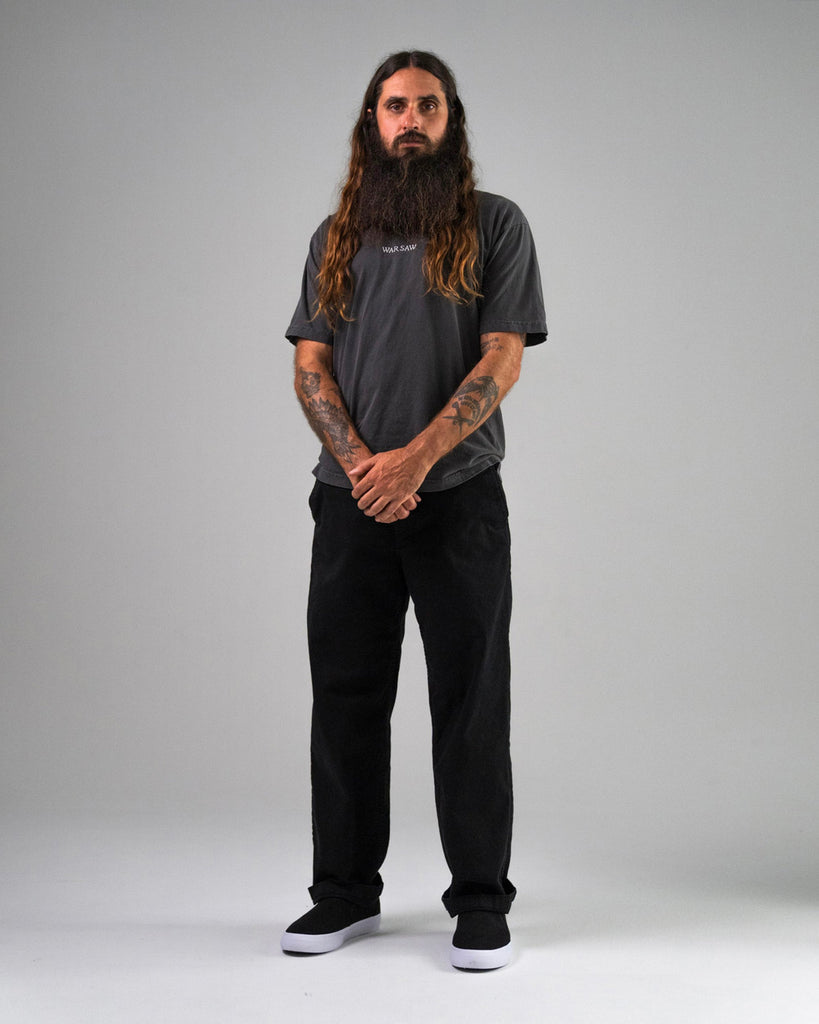 A man with long hair and a beard stands against a plain gray background, wearing a dark T-shirt, black pants, and WARSAW LTD STRANGER SLIP-ON shoes in black/white synthetic with a minimalistic skate-inspired silhouette.