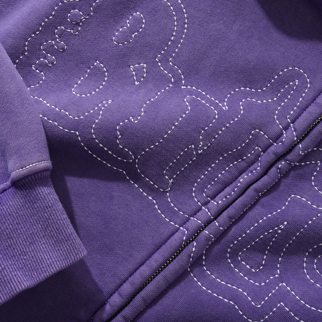 Close-up of a washed purple BUTTER BREAKDOWN ZIP-THRU HOODIE from Butter Goods, showcasing visible stitching outlining a stylized skull and text design, featuring direct embroidery artwork.