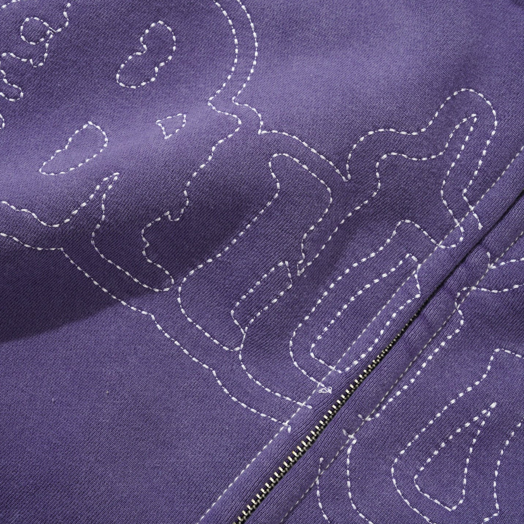 Close-up of a washed purple fabric from the BUTTER BREAKDOWN ZIP-THRU HOODIE WASHED PURPLE by Butter Goods, featuring a white stitched outline of a skull and crossbones design, with a visible metal zipper running through the image. The direct embroidery artwork is showcased beautifully on this Cotton/Polyester Blend.