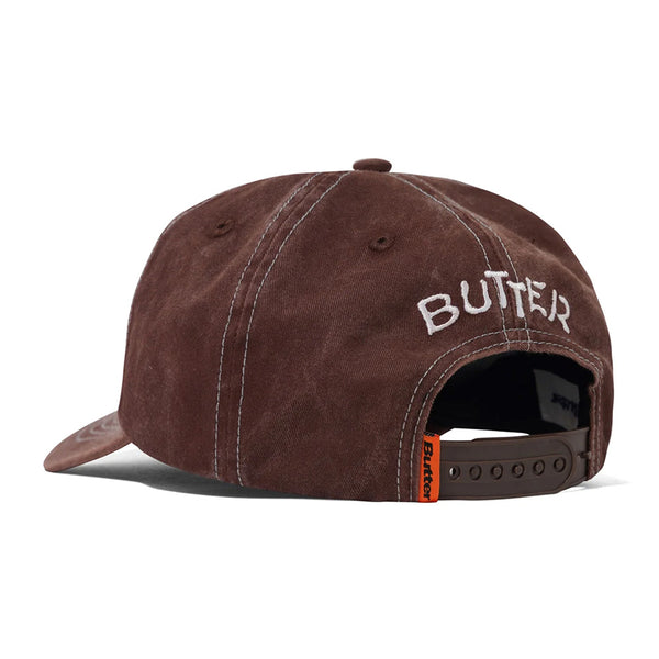 The Butter Goods BUTTER CANINE 5 PANEL CAP WASHED BROWN is a brown 5-panel cap shown from the back, with "BUTTER" embroidered above the snapback closure and an orange tag on the strap.