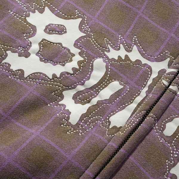 Close-up of the BUTTER ERUPTION ZIP-THRU HOODIE PLAID by Butter Goods, featuring purple and brown plaid fabric, a white abstract design, visible stitching, and a vertical zipper.