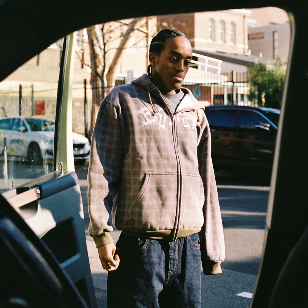 A person wearing the Butter Goods BUTTER ERUPTION ZIP-THRU HOODIE PLAID and dark jeans stands on a city street, visible through a vehicle's open door.