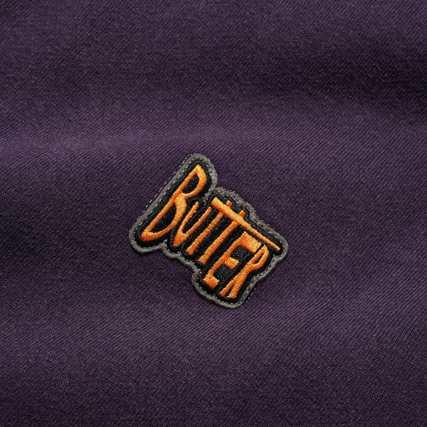 A close-up of the Butter Goods BUTTER SKETCH ZIP-THRU HOODIE EGGPLANT shows its purple mineral wash fabric with an orange and black "Butter" embroidered patch.