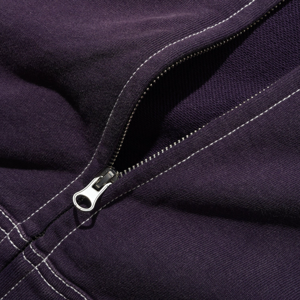A close-up of the partially unzipped silver zipper on dark purple fabric with white stitching highlights the detail of the Butter Goods BUTTER SKETCH ZIP-THRU HOODIE EGGPLANT.