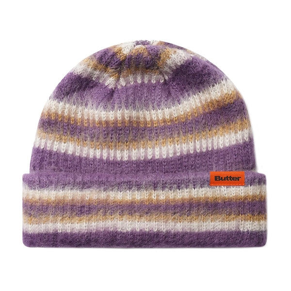 The Butter Goods BUTTER SWIPE BEANIE PURPLE features purple, white, and tan jacquard stripes, a folded cuff, and an orange rectangular "Butter" label on the front.