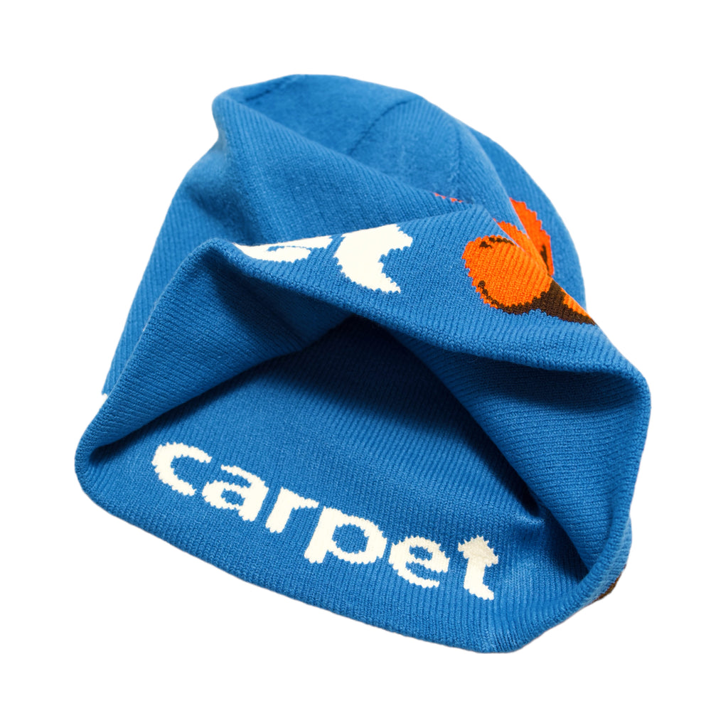 The CARPET CO. BACTERIA REVERSIBLE BEANIE BLUE from Carpet Co. is a blue knitted hat adorned with the word "carpet" and a vibrant design in orange, white, and black. Its slightly crumpled appearance adds to its unique charm and authentic style.