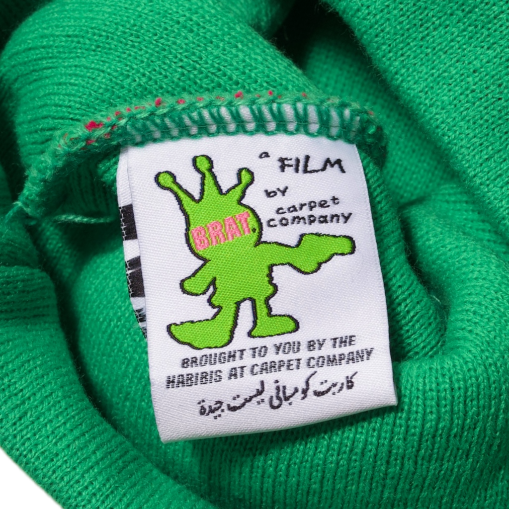Clothing label featuring a green monster graphic with the logo "a film by Carpet Co." on a textured green fabric background. (Product: Carpet Co. Brat Logo No Fold Beanie Green)