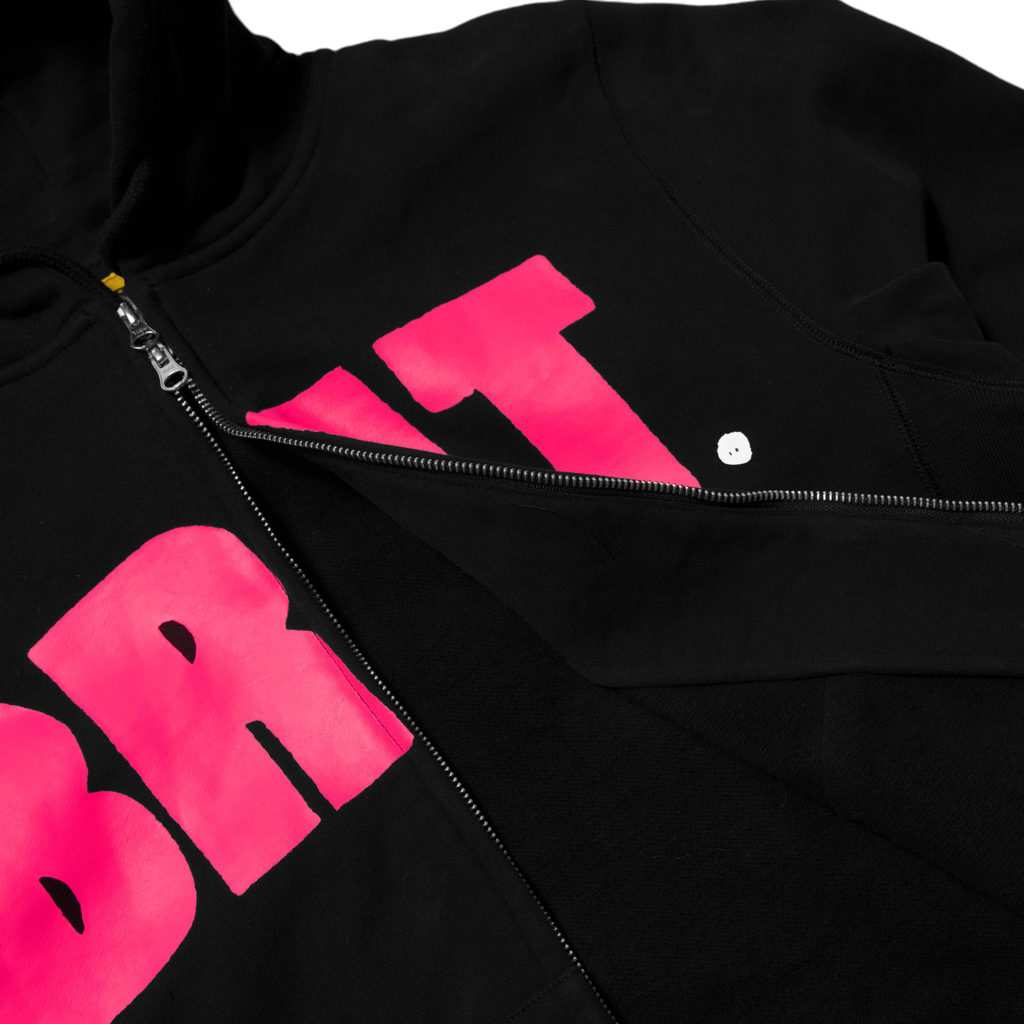 Carpet Co. black zip hoodie with vibrant pink "brit" lettering, partially zipped with a visible white snap button at the neckline.