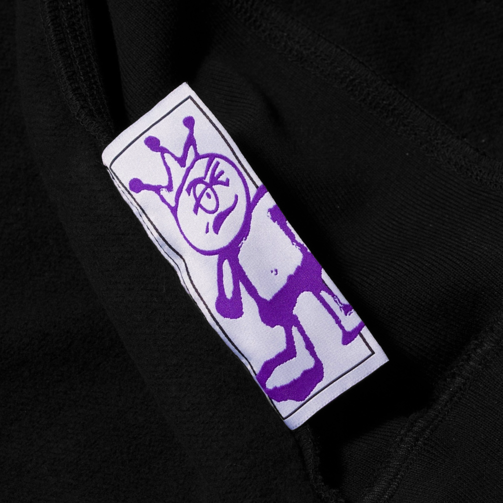 A purple, hand-drawn character on a white clothing label sewn inside a Carpet Co. Brat Logo Zip Hoodie Black.