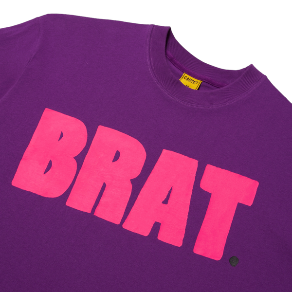 A close-up of a Carpet Co. brat tee in purple with the word "brat" screen printed in bold pink letters across the chest.