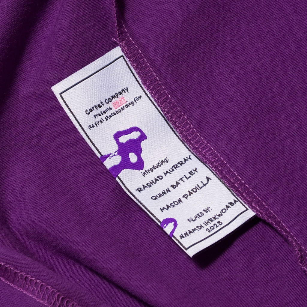 Close-up of a Carpet Co. BRAT TEE PURPLE garment label on purple fabric showing care instructions and fabric composition details.