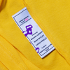 Close-up of a clothing label on a yellow fabric listing designer names and the brand 'Carpet Co.,' with garment details of the CARPET CO. BRAT TEE LEMON.