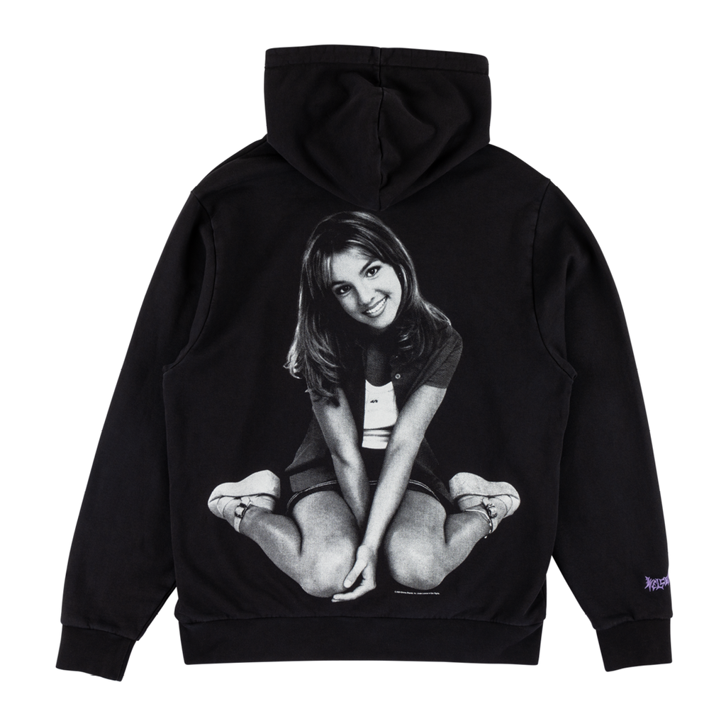 The WELCOME X BRITNEY FLOWER PIGMENT-DYED HOODIE BLACK features a black and white graphic of a woman sitting cross-legged and smiling at the camera, screen printed on the back. It also boasts a purple WELCOME logo on the left sleeve.