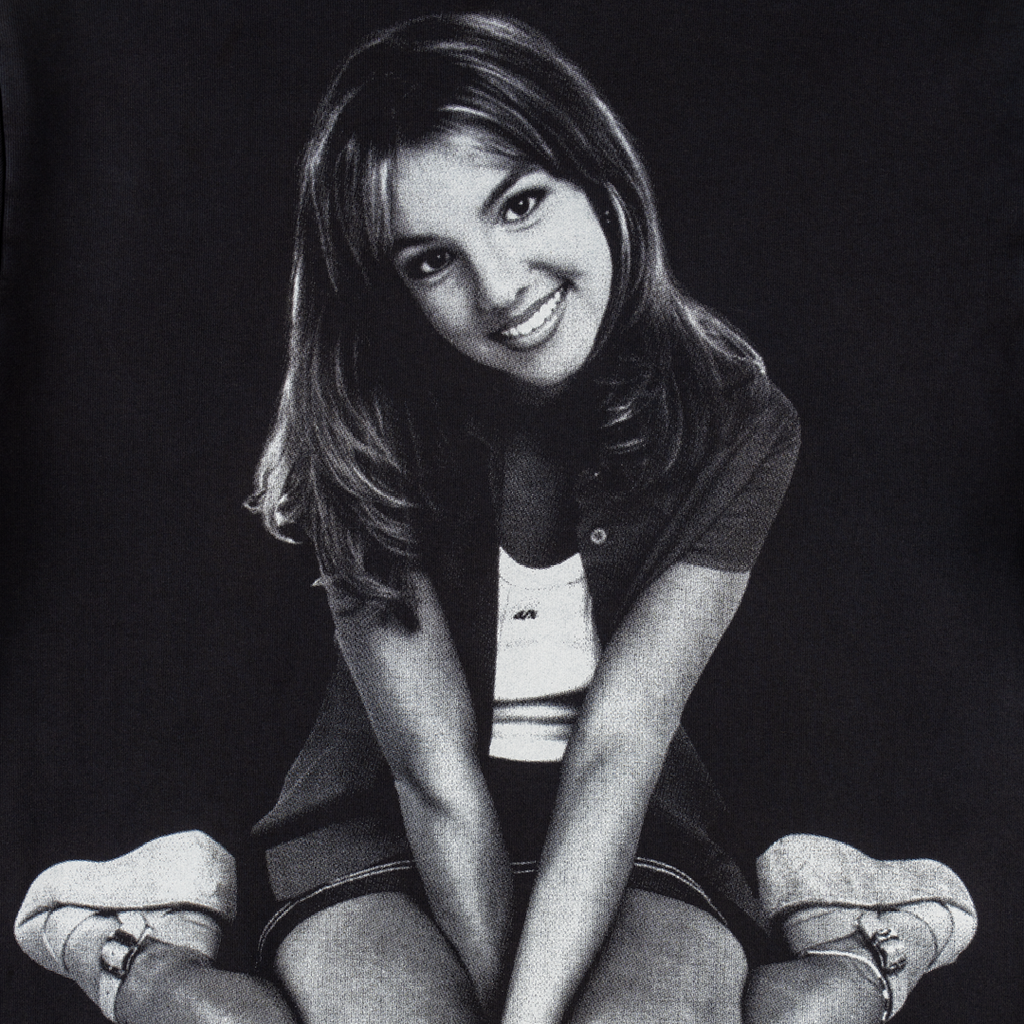 Black-and-white photo of a young woman sitting on the floor with her legs crossed and hands on her knees, smiling at the camera in a WELCOME X BRITNEY Flower Pigment-Dyed Hoodie in black.