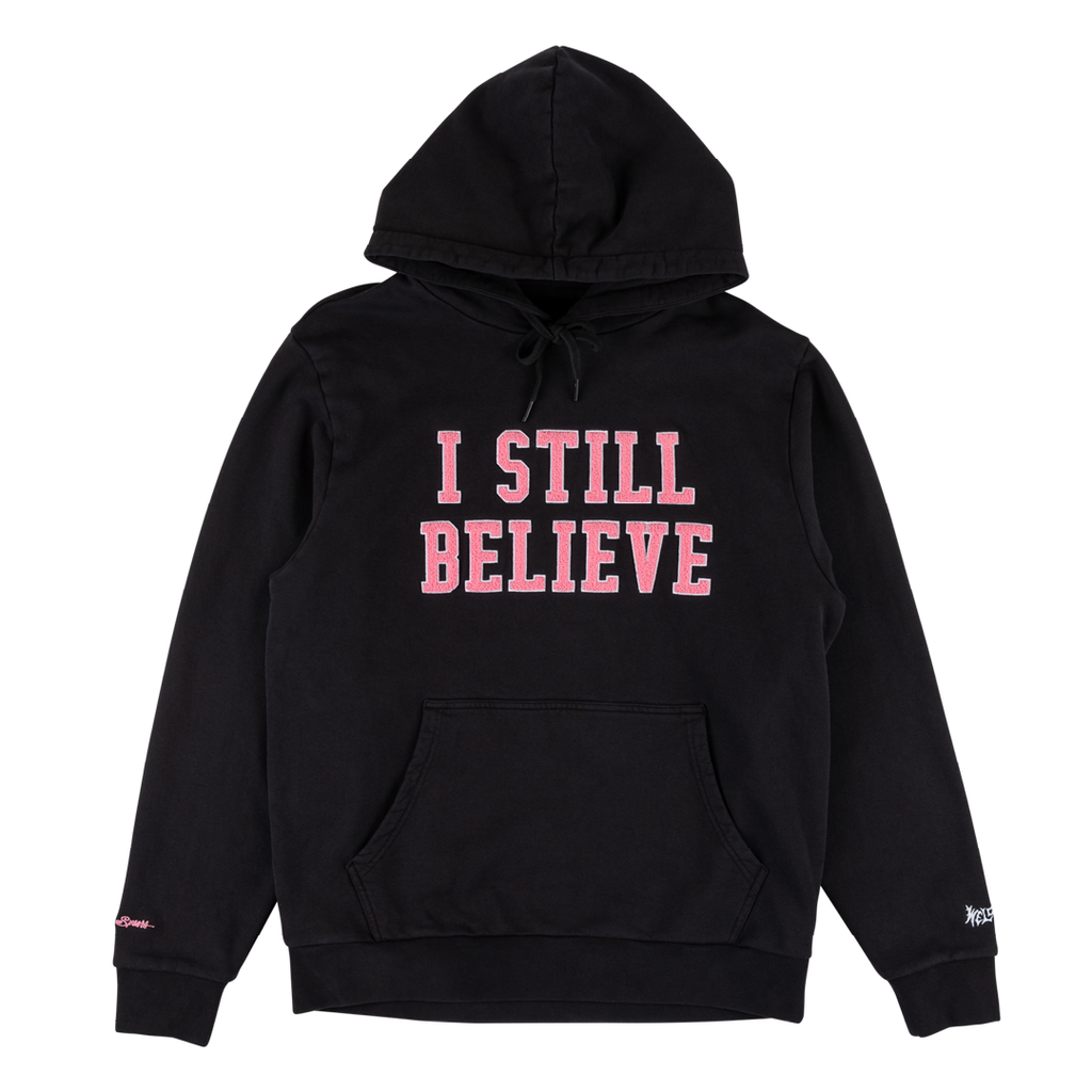 The WELCOME X BRITNEY BELIEVE Pigment-Dyed Hoodie in Black showcases the bold pink capitalized text "I STILL BELIEVE" screen printed on the front. This stylish and practical hoodie from WELCOME features a front pocket and a drawstring hood.