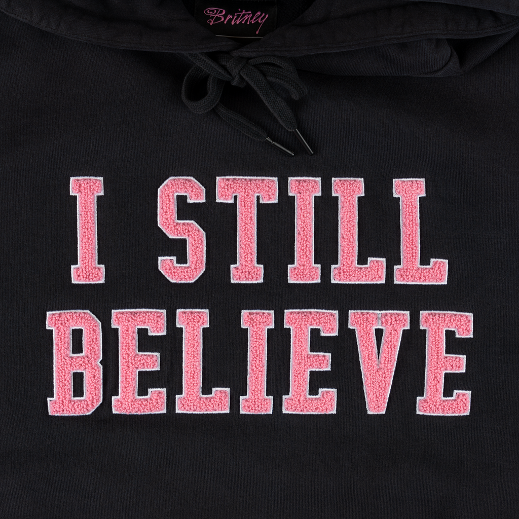 Close-up of a WELCOME X Britney Believe Pigment-Dyed Hoodie Black, featuring "I STILL BELIEVE" in large pink and white letters screen printed on the front.