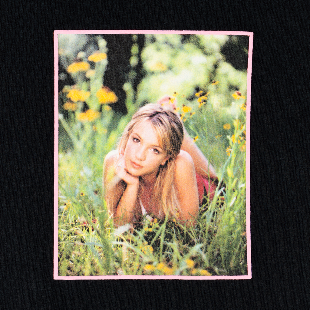 A person lying in a field of grass and yellow flowers, resting on their elbows and looking at the camera, wearing a Welcome X Britney Daisies Tee in black. The image is framed with a pink border.