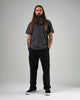 A man with long hair and a beard stands facing forward, wearing a dark t-shirt, black pants, and WARSAW LTD CHAPTER MID | BLACK / WHITE SYNTHETIC skate footwear, his hands in his pockets against a plain background.