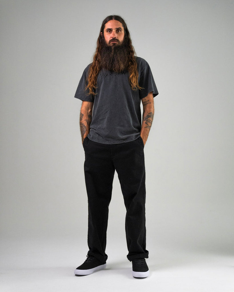 A man with long hair and a beard stands facing forward, wearing a dark t-shirt, black pants, and WARSAW LTD CHAPTER MID | BLACK / WHITE SYNTHETIC skate footwear, his hands in his pockets against a plain background.