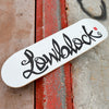 A skateboard featuring "CHOCOLATE" in stylized black lettering on a white deck rests on a textured concrete surface, reflecting the innovative spirit of future builds from the CHOCOLATE X BLUETILE X LOWBLOCK FLIP THE SCRIPT series.