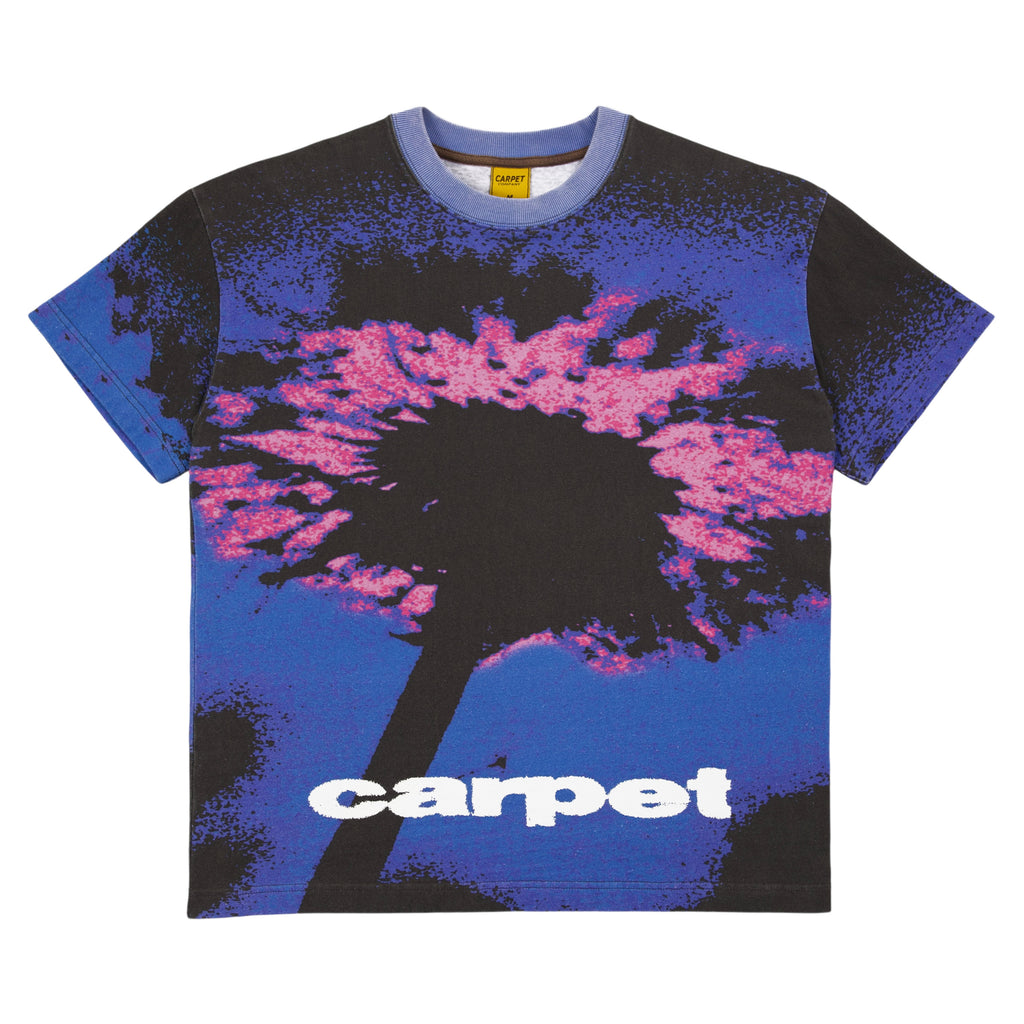 Introducing the Carpet Dandelion Tee Blue by Carpet Co.: a blue shirt featuring a striking graphic of a dandelion in pink and blue tones, screen printed to perfection. This unique design includes the word "carpet" subtly at the bottom, adding an artistic touch to your wardrobe.