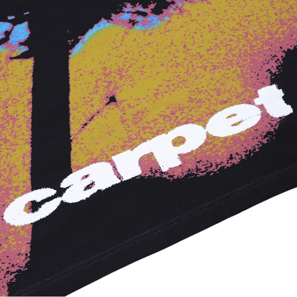 The image features a detailed view of black fabric adorned with a pixelated yellow and pink pattern, resembling the lively design of the CARPET DANDELION TEE YELLOW by Carpet Co. The word "carpet" prominently appears in bold white letters, drawing attention amidst the complex design.