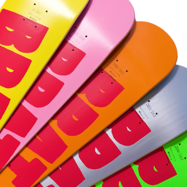 Collection of colorful Carpet Company Brat Logo snowboards with face graphics lined up, varying from pink to orange and grey, each screen printed.