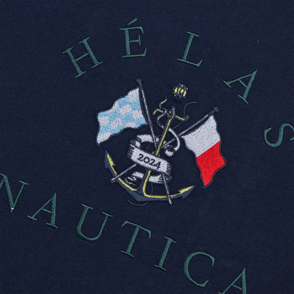 Close-up of an embroidered emblem on a HELAS X NAUTICA THIBAULT TEE DARK NAVY, featuring an anchor with crossed blue and white checkered and red, white, and blue flags, the word "HÉLAS NAUTICA" and the year "2021".