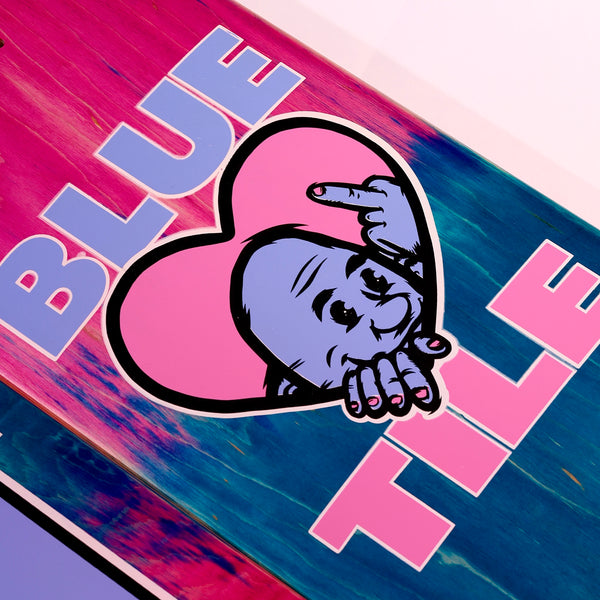 The BLUETILE F.U. DECK by Bluetile Skateboards features a pink and blue gradient background with the text "BLUE TILE" and a cartoon character inside a pink heart, pointing with one finger. This deck showcases Jay Croft's artwork, resulting in an eye-catching design.