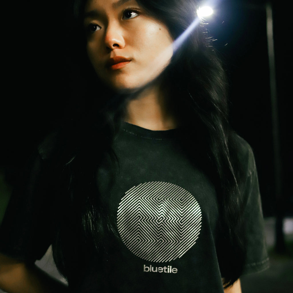 A person with long black hair stands outdoors at night, showcasing a black BLUETILE "Crazy" Silver Foil Stone Washed Tee, featuring a circular design and the word "BLUETILE," reflecting the bold style of Bluetile Skateboards.