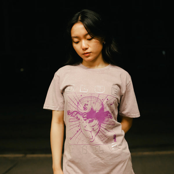 In the dim light, a person stands wearing Bluetile Skateboards' BLUETILE "WOKE" Silver Foil Stone Washed Tee in Orchid, featuring a subtle reflective 3M print that catches your eye.