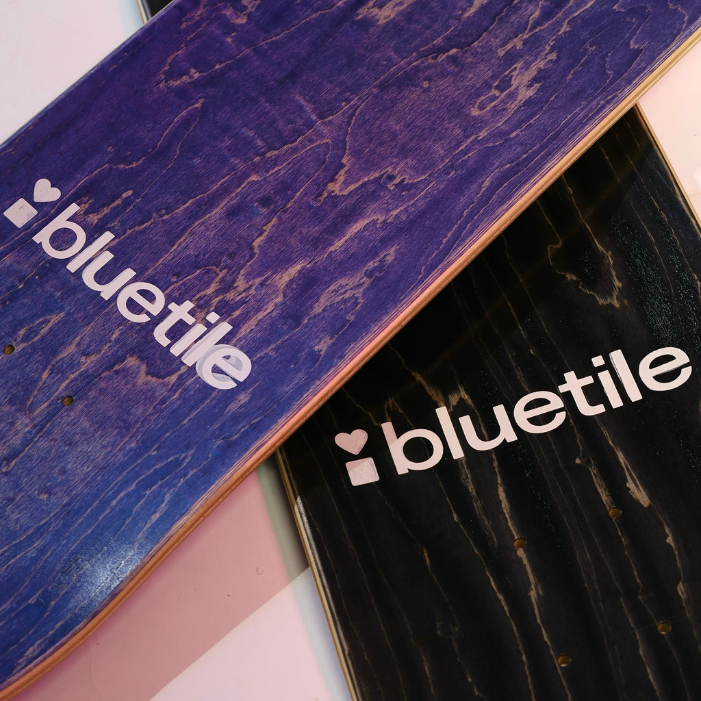 Close-up of two BLUETILE W.T.F. DECK skateboard decks by Bluetile Skateboards; one is blue and the other is black, both featuring Jay Croft artwork with the word "bluetile" in white and a heart logo.