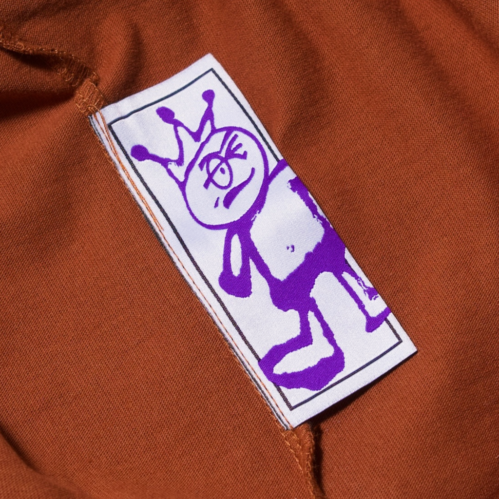 Alt text: a purple doodle of a stick figure with a crown, hand screen printed on a white rectangular label sewn onto Carpet Co. "Kid" Tee Brown fabric.